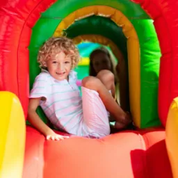 Uśmiechnięty chłopiec z kręconymi blond włosami siedzi wewnątrz kolorowego, dmuchanego tunelu z czerwonymi, żółtymi i zielonymi elementami, w tle widoczna częściowo zamazana postać.