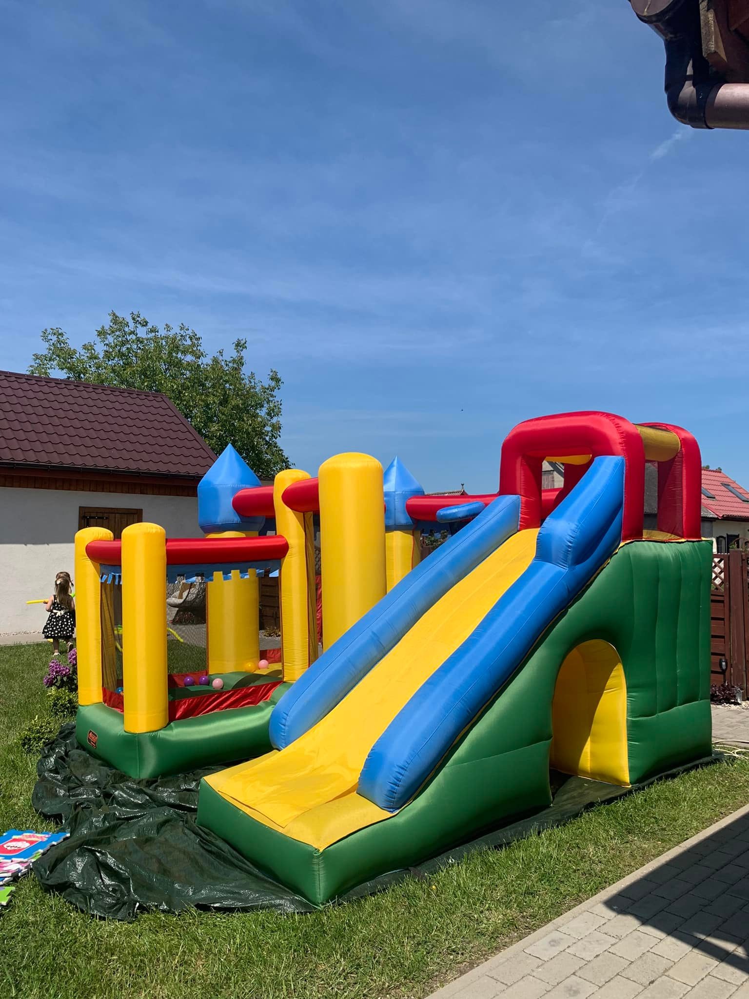 Kolorowy, dmuchany plac zabaw z zamkiem i zjeżdżalnią na trawniku w słoneczny dzień, z widoczną dziewczynką w tle.