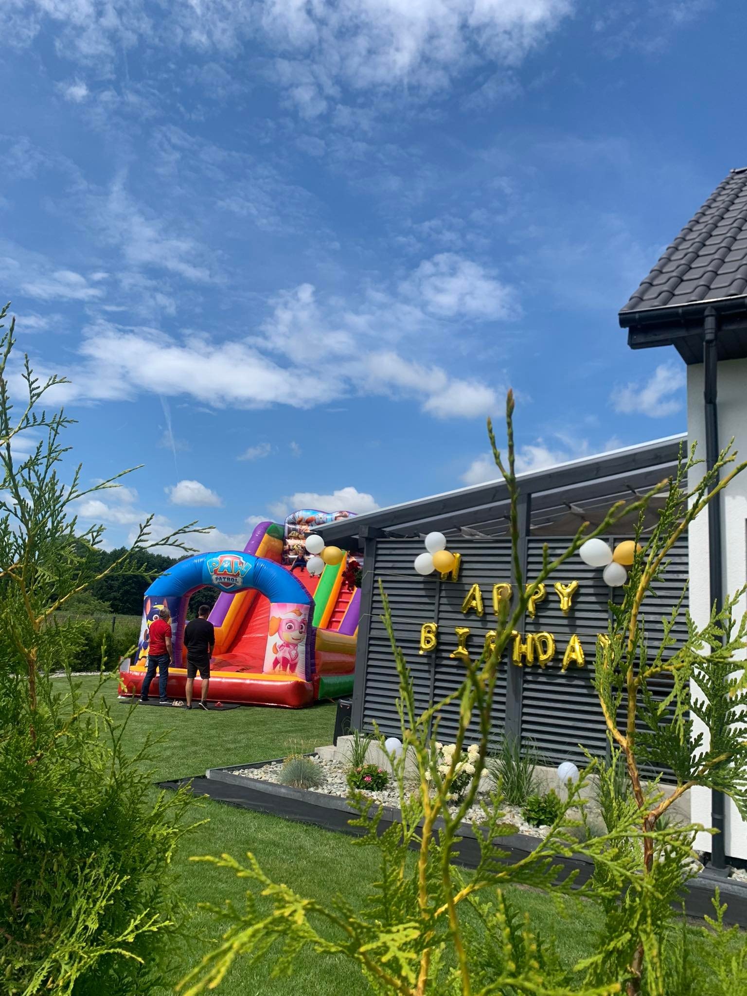 Kolorowy dmuchany zamek z motywem Psi Patrol na trawniku, z dekoracją 'Happy Birthday' z balonów, widoczny przez iglaki