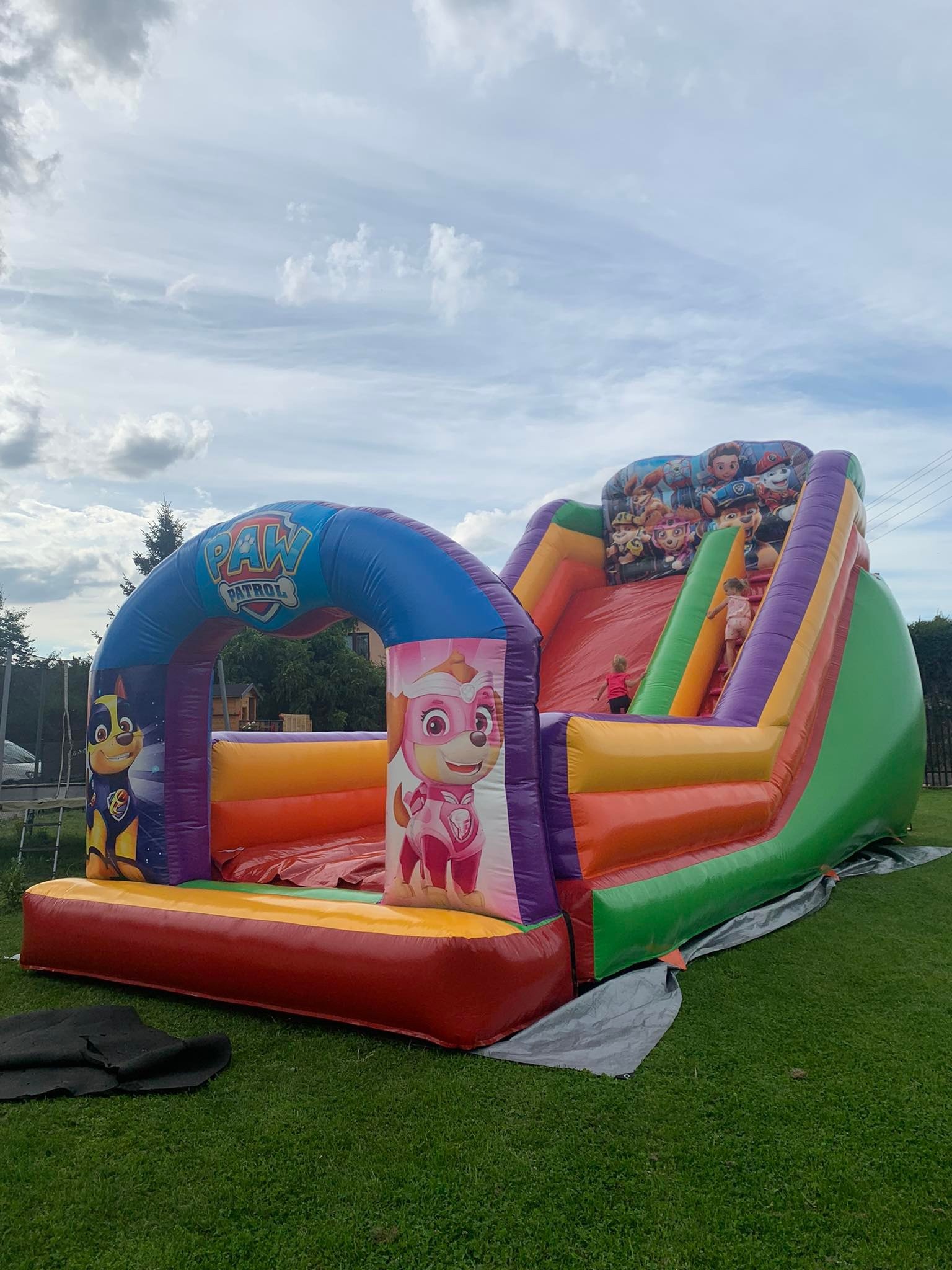 Kolorowa dmuchana zjeżdżalnia z motywem bajki Psi Patrol, ustawiona na trawniku, z dwojgiem dzieci w trakcie zabawy.