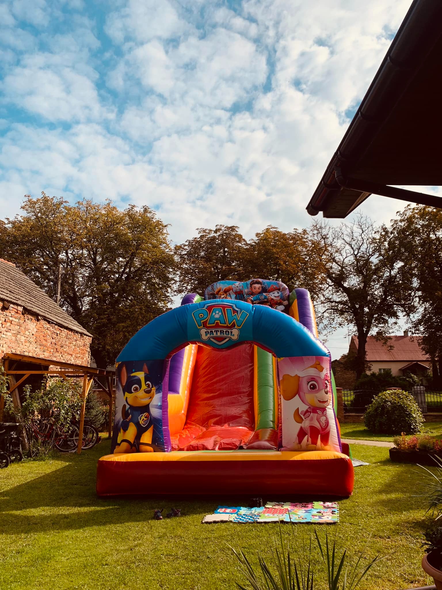 Kolorowy dmuchany zamek ze zjeżdżalnią, motyw Psi Patrol, ustawiony na zielonym trawniku w ogrodzie, w tle budynek z czerwonej cegły i niebieskie niebo z chmurami.