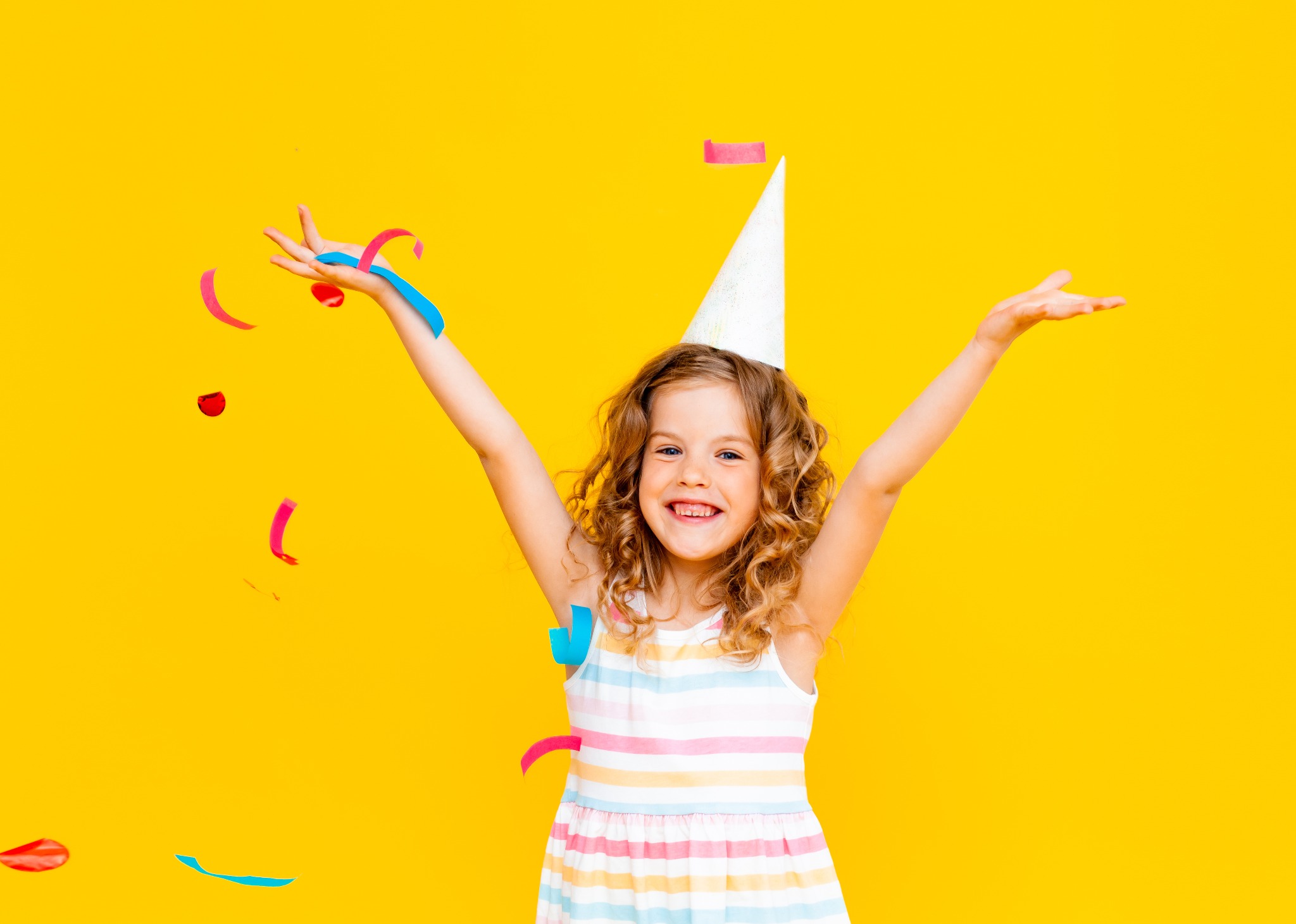 Uśmiechnięta dziewczynka w stroju w paski i papierowej czapeczce, z rozrzuconymi kolorowymi serpentynami na jaskrawożółtym tle.