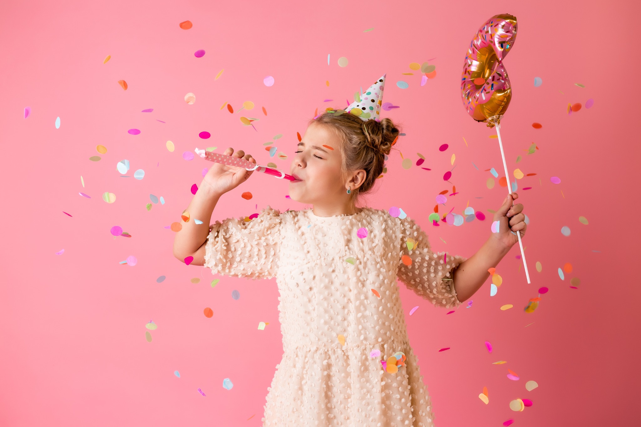 Dziewczynka w sukience i czapeczce urodzinowej dmucha w serpentynę, trzymając balon w kształcie pączka na różowym tle, otoczona konfetti.