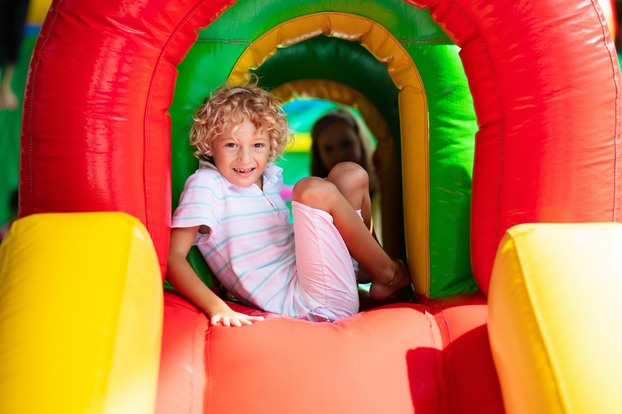 Uśmiechnięty chłopiec z kręconymi blond włosami siedzi wewnątrz kolorowego, dmuchanego tunelu z czerwonymi, żółtymi i zielonymi elementami, w tle widoczna częściowo zamazana postać.