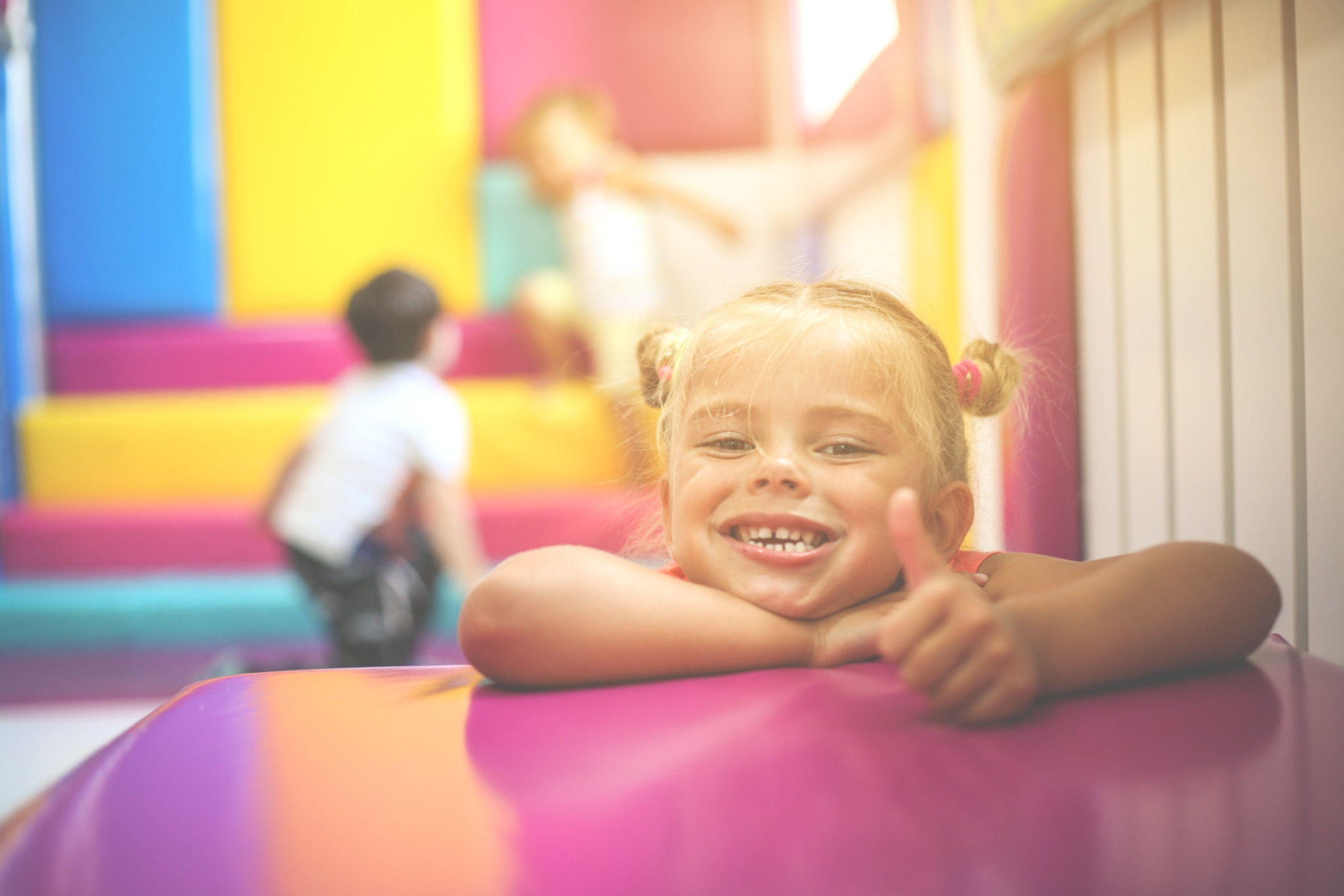 Uśmiechnięta dziewczynka z blond włosami spiętymi w dwa kucyki, opiera się o różowy stół, pokazując kciuk w górę na tle kolorowego placu zabaw w Szczecinie.