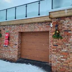 Murowana elewacja budynku z garażem i szklaną balustradą na dachu, udekorowana świątecznie figurką św. Mikołaja na linie i wieńcem z szyszkami, w tle pada śnieg.