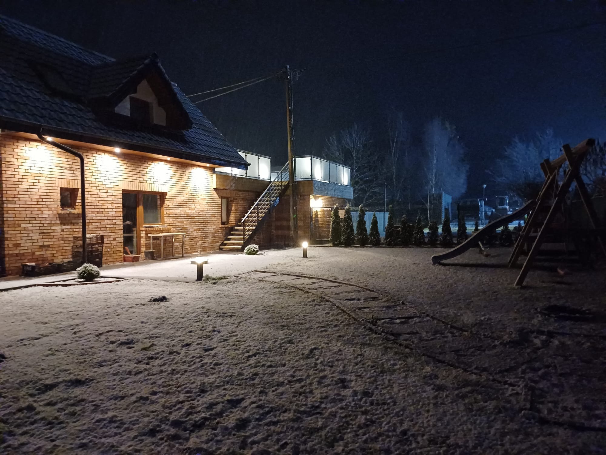 Oświetlona nocą elewacja domu z czerwonej cegły z zewnętrznymi schodami prowadzącymi na taras, pokryta delikatnym śniegiem.