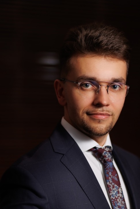 Portret młodego mężczyzny w garniturze i okularach, z delikatnym uśmiechem, na ciemnym, rozmytym tle. Elegancki wygląd, profesjonalny wizerunek.