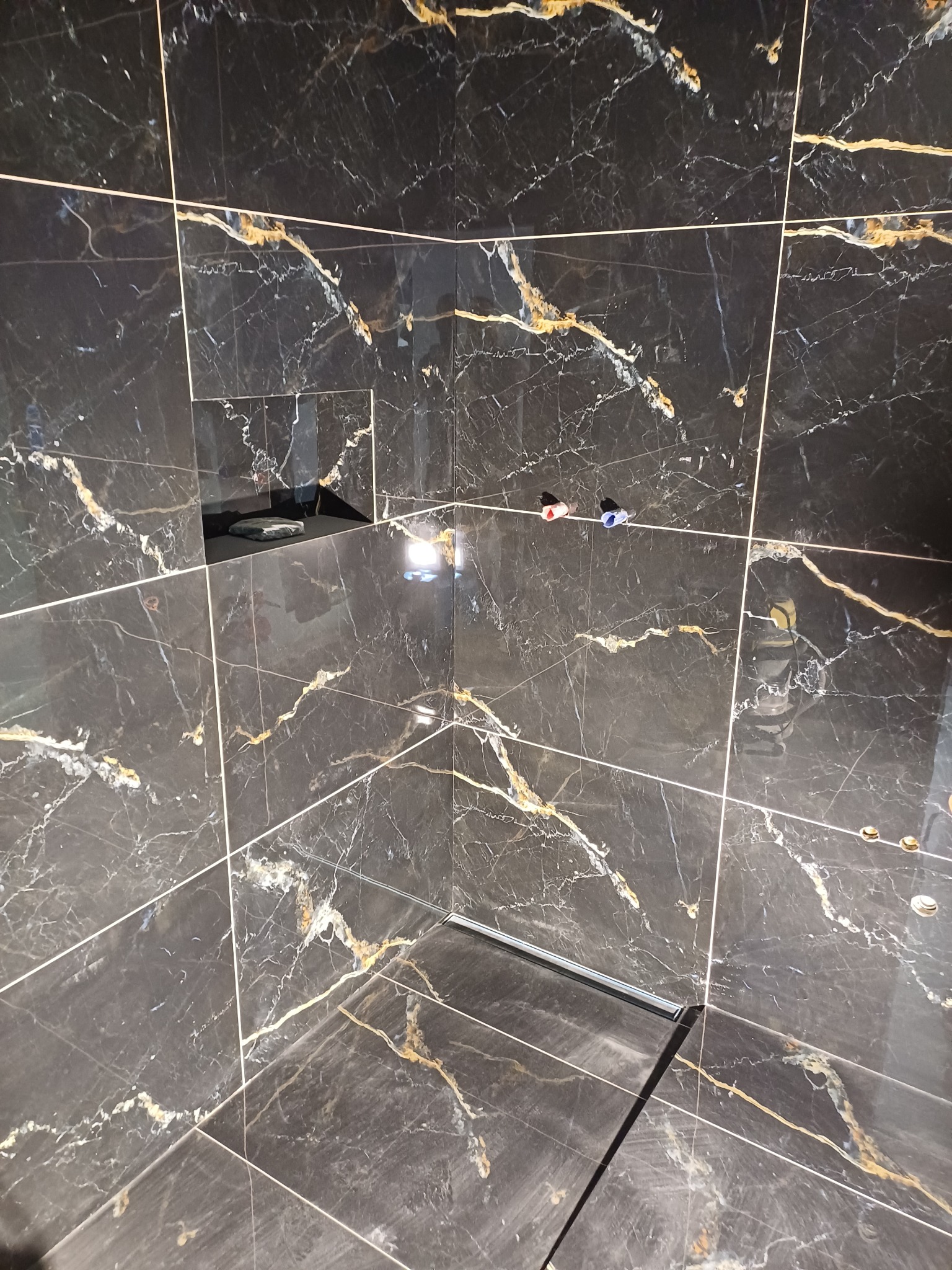 Kabina prysznicowa wyłożona czarnymi płytkami imitującymi marmur ze złotymi żyłami. Widoczny odpływ liniowy i elementy instalacji wodnej.