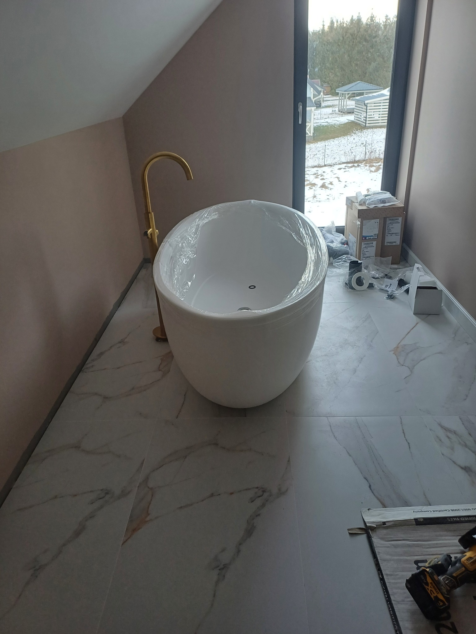 Nowoczesna, wolnostojąca wanna owalna z złotą armaturą w łazience z oknem. Na podłodze płytki imitujące marmur. Widoczne narzędzia i materiały montażowe.