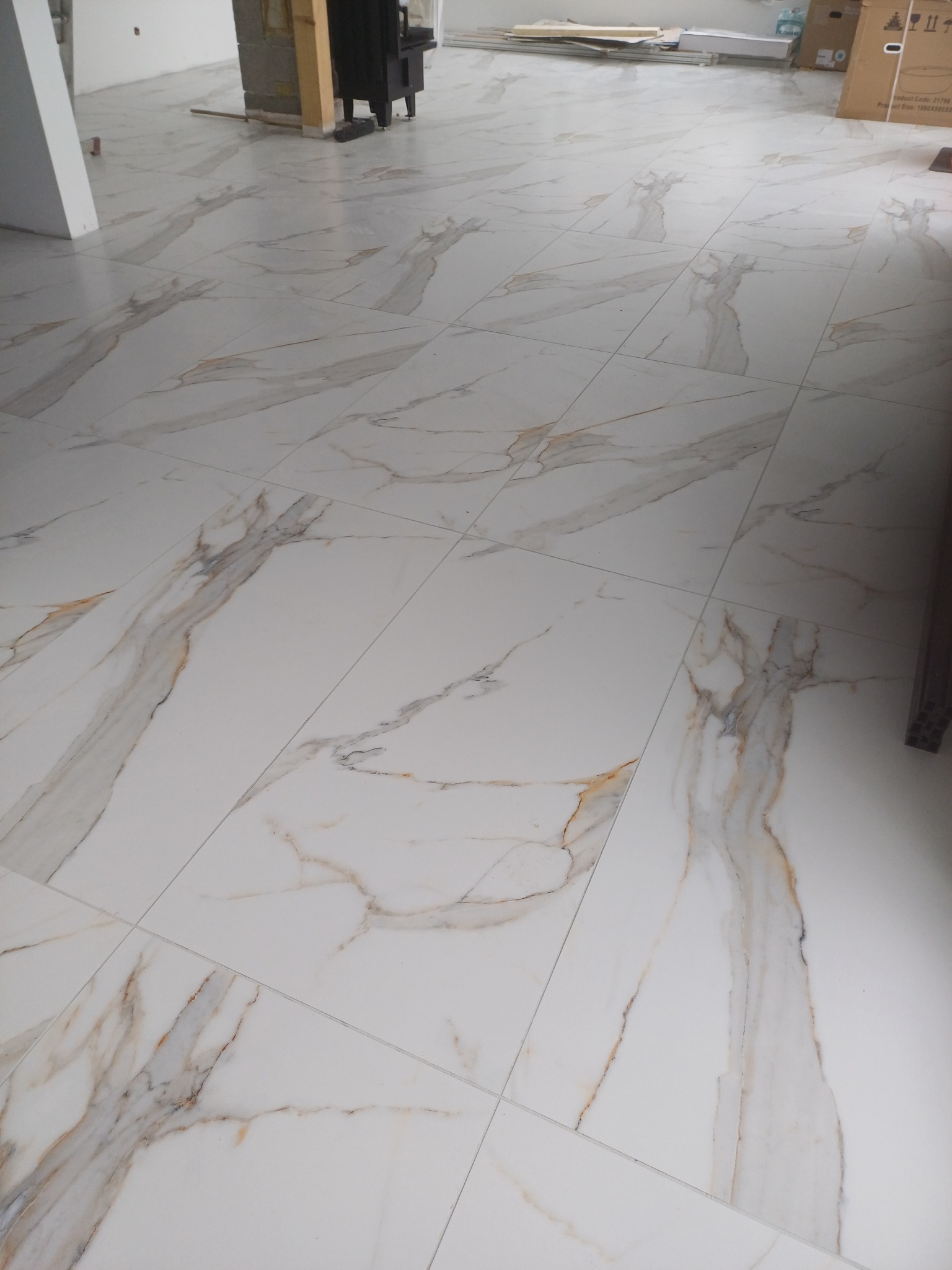 Ułożone, białe płytki podłogowe z marmurkowym wzorem w odcieniach szarości i złota. Widoczne fugi. Częściowo widoczne elementy wyposażenia wnętrza w tle.