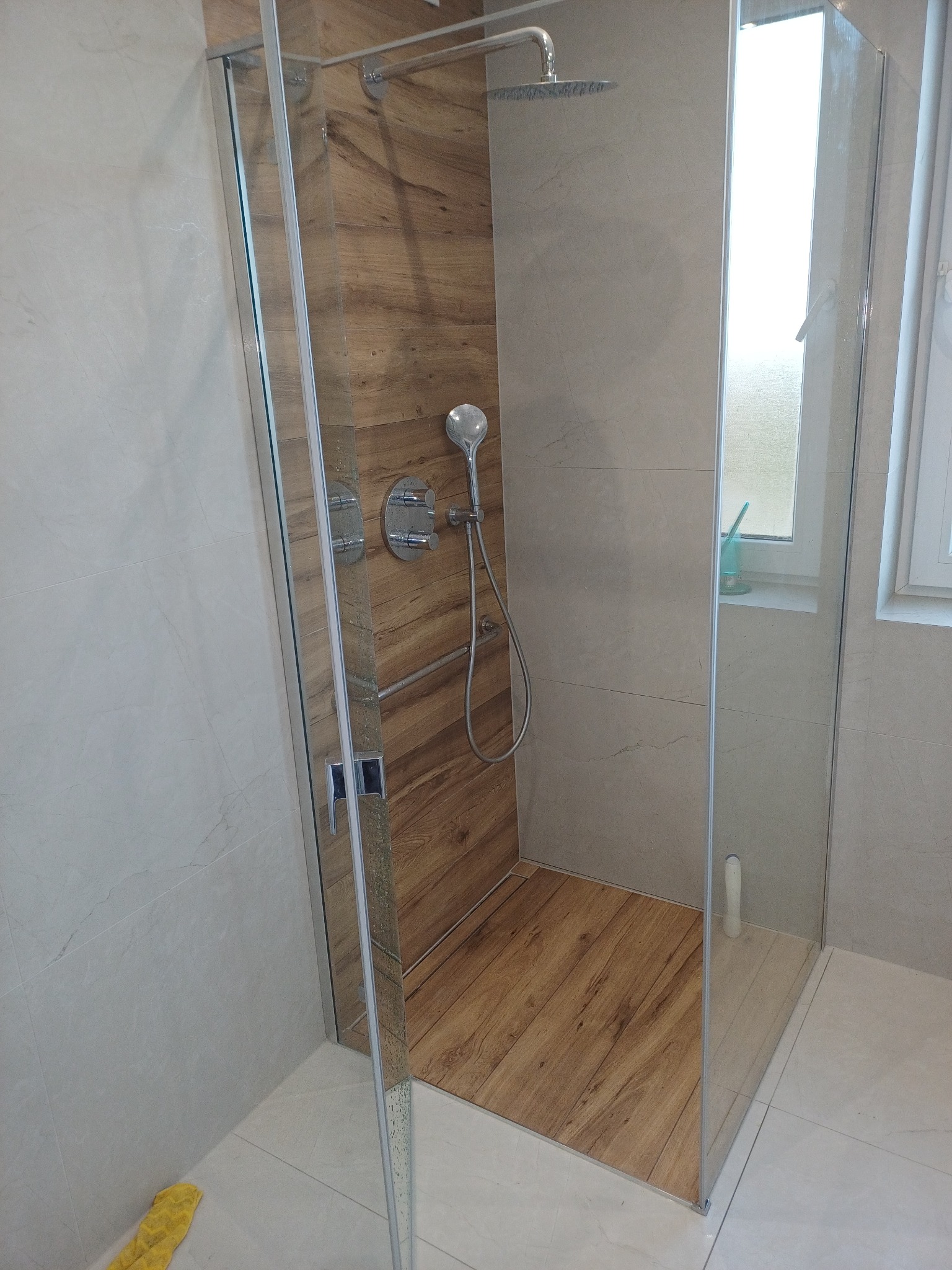 Kabina prysznicowa walk-in z drewnopodobną okładziną wewnątrz, szklane ściany, deszczownica i bateria termostatyczna. Widok na okno z akcesoriami łazienkowymi.