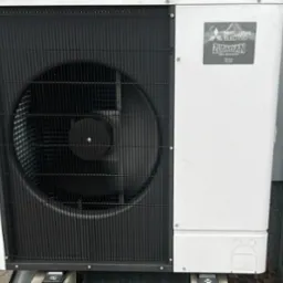 Zewnętrzna jednostka pompy ciepła Mitsubishi Zubadan R32, widoczna czarna kratka wentylatora i biała obudowa, zamontowana przy ścianie budynku.