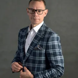 Portret mężczyzny w okularach i kraciastej marynarce, zapinającego guzik, na szarym tle.