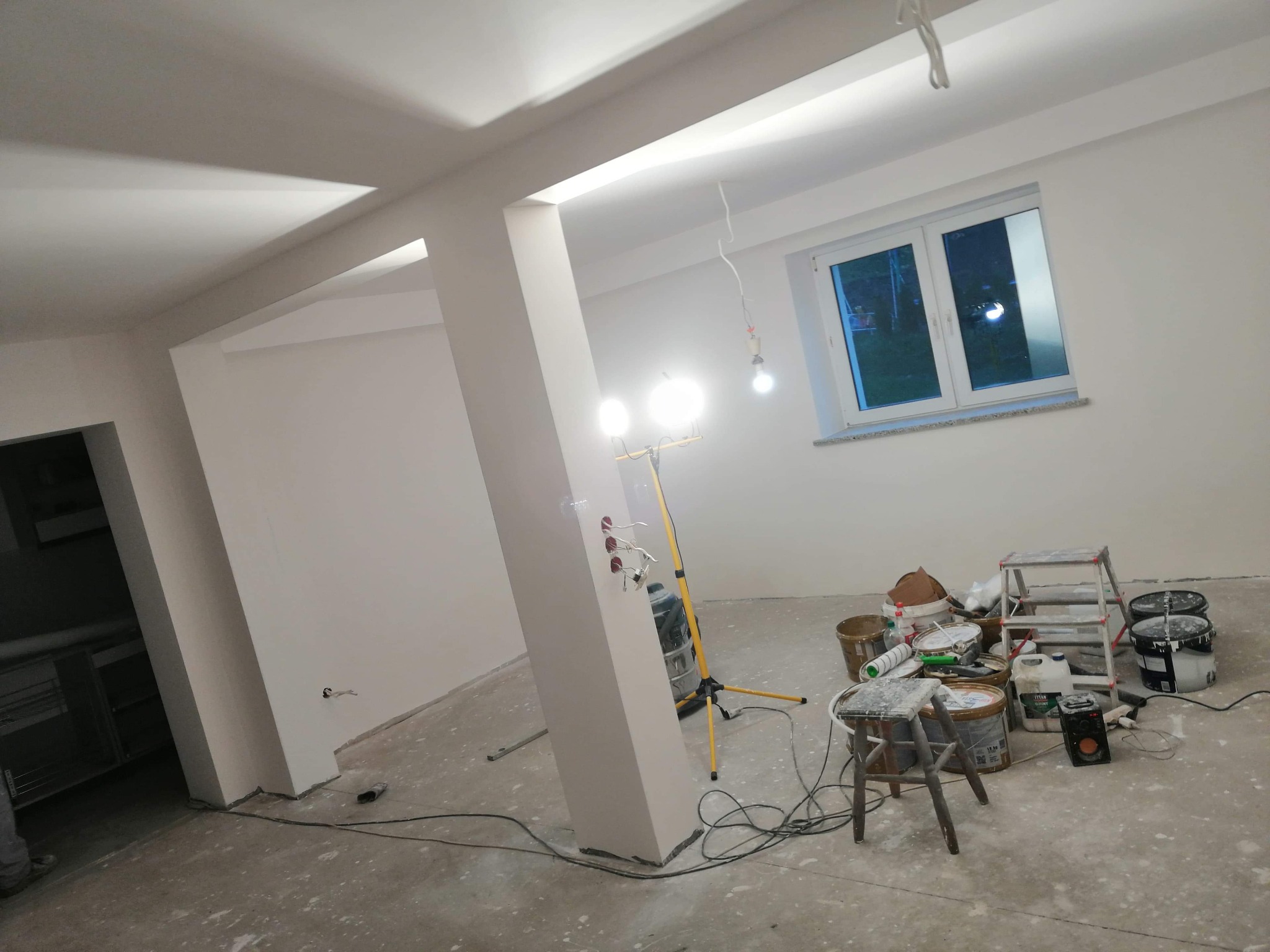 Wnętrze w trakcie prac wykończeniowych: świeżo pomalowane ściany na biało, widoczne narzędzia malarskie, drabina, wiaderka z farbą oraz lampa budowlana oświetlająca filar z gniazdkami elektrycznymi.