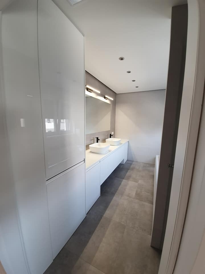 Nowoczesna łazienka z białymi szafkami, dwoma umywalkami nablatowymi i szarą podłogą z płytek. Oświetlenie punktowe w suficie i podłużne nad lustrem.