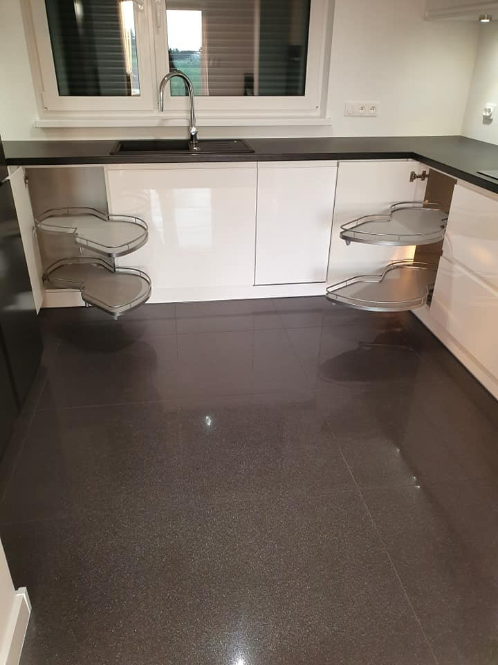 Nowoczesna kuchnia z błyszczącymi, białymi szafkami, czarnym blatem i ciemną podłogą; otwarte szafki narożne z obrotowymi półkami.