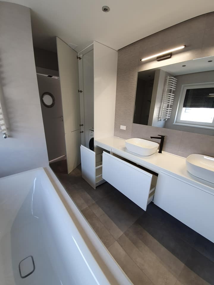 Nowoczesna łazienka z białą wanną, podwójną umywalką nablatową, szafkami z otwartymi szufladami i ukrytą pralką za białymi drzwiami w zabudowie.