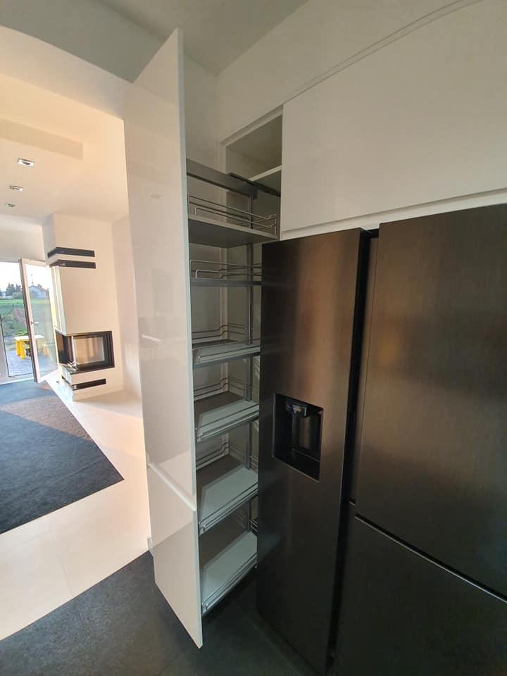 Nowoczesna kuchnia z białymi szafkami na wysoki połysk i wysuwanym systemem cargo, obok czarna lodówka z dystrybutorem wody.