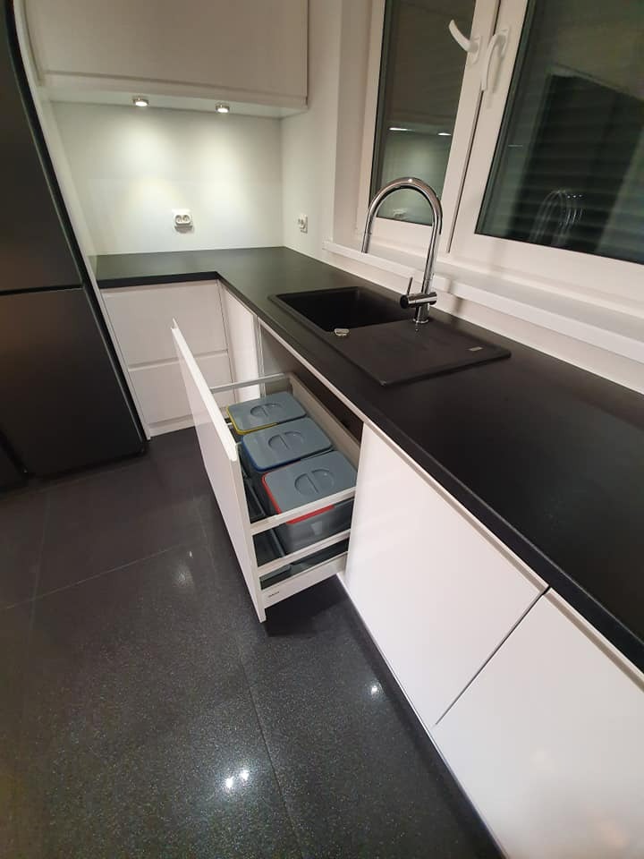 Nowoczesna kuchnia z czarnym blatem i białymi szafkami, wysuwany kosz na śmieci z trzema pojemnikami, widok na okno z chromowaną baterią.