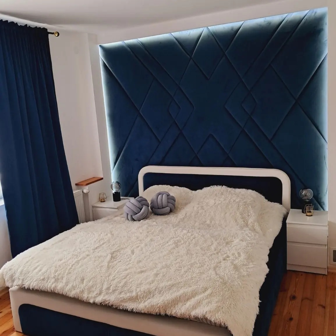 Niebieski, tapicerowany panel ścienny z geometrycznym wzorem, podświetlony od tyłu, nad łóżkiem z puszystą, białą narzutą i dekoracyjnymi poduszkami w kształcie węzłów.