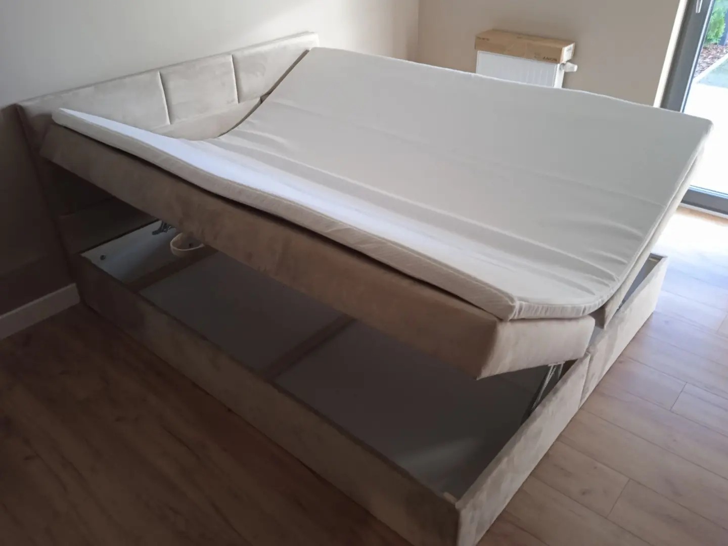 Beżowe łóżko tapicerowane z podnoszonym stelażem i pojemnikiem na pościel, widoczne mechanizmy podnoszenia.