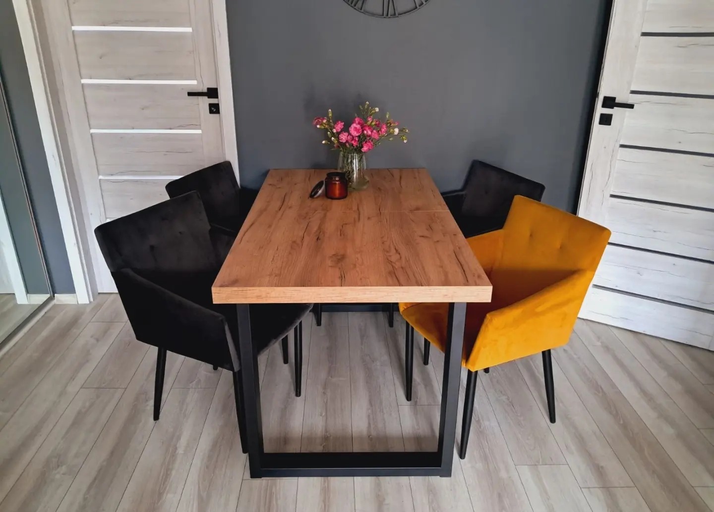 Drewniany stół z czarnymi nogami otoczony aksamitnymi krzesłami w kolorze czarnym i musztardowym, na blacie wazon z różowymi kwiatami i świeca.