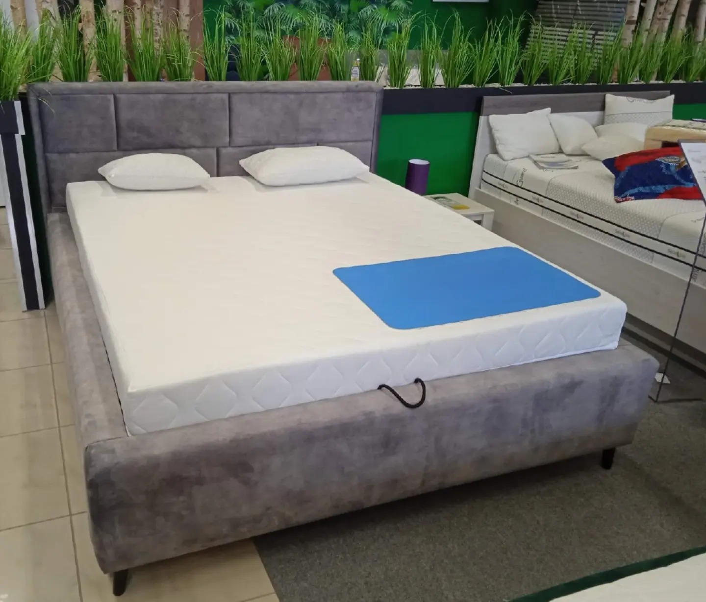 Szare łóżko tapicerowane z pikowanym zagłówkiem i białym materacem, widoczna niebieska podkładka ochronna na materacu oraz rośliny w tle.