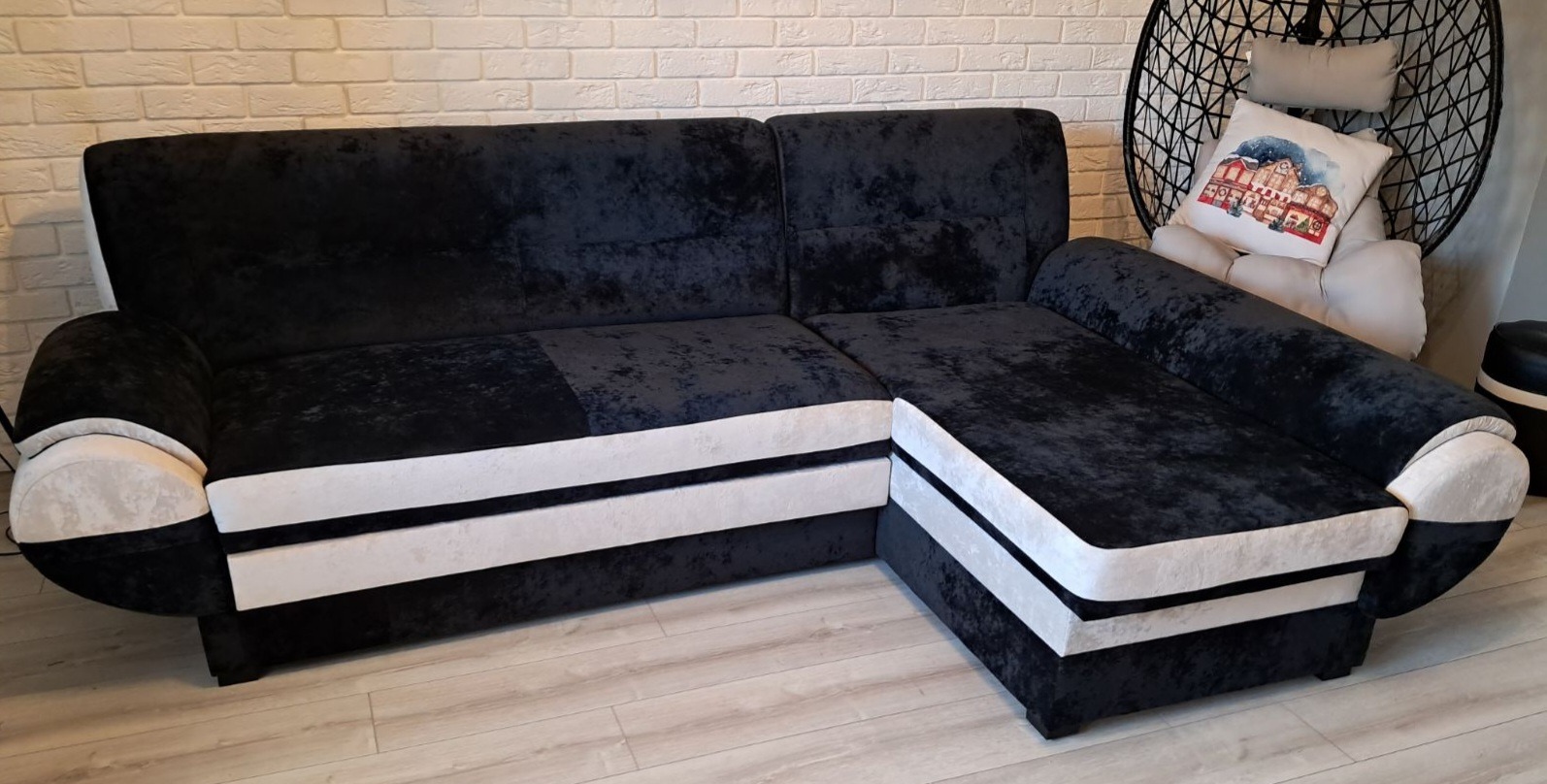 Czarno-biały narożnik z welurową tapicerką i jasnymi bokami, na tle ściany z białej cegły i wiszącego fotela.