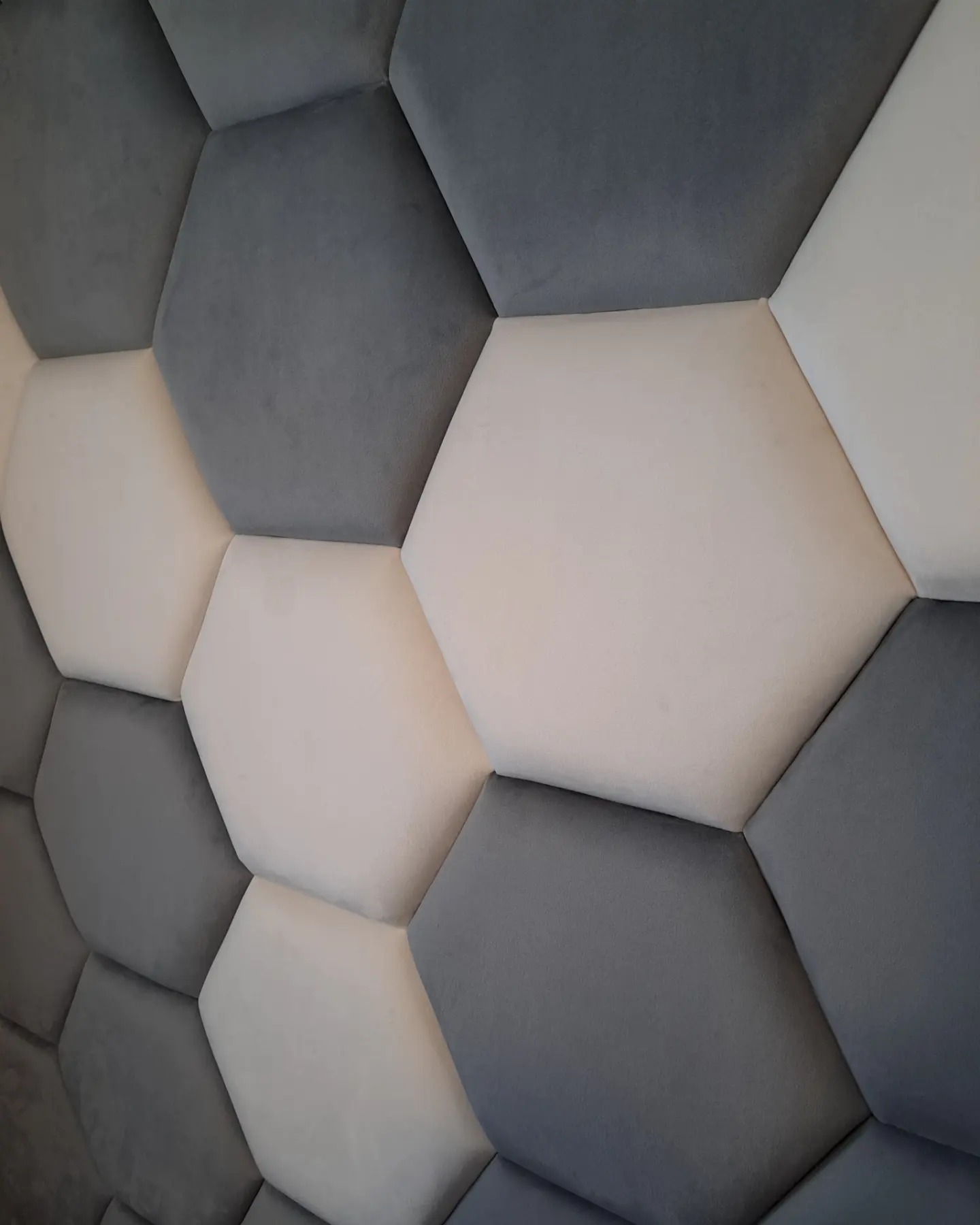 Dekoracyjny panel ścienny z miękkich, tapicerowanych heksagonów w odcieniach szarości i beżu, tworzących trójwymiarową teksturę.
