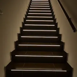 Schody z ciemnymi płytkami i podświetleniem LED na każdym stopniu, widok z dołu, poręcz po prawej stronie.