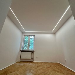 RENONEUF - Pusty pokój po remoncie: parkiet w jodełkę, okno z widokiem na zieleń i nowoczesne oświetlenie LED w suficie, tworzące geometryczny wzór.