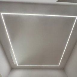 RENONEUF - Minimalistyczne oświetlenie sufitowe LED w kształcie kwadratu, w białym pomieszczeniu z widocznym fragmentem okna i drzwi.