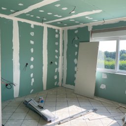 RENONEUF - Pokój w trakcie remontu: zielone płyty gipsowo-kartonowe na ścianach, częściowo zaszpachlowane, widoczne przewody, poziomica, drabina. Przygotowanie do dalszych prac.