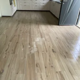 Ułożona podłoga z paneli imitujących drewno w jasnej kuchni z białymi szafkami i czarną mozaiką nad blatem. Widoczna nowoczesna, szara rama drzwi.