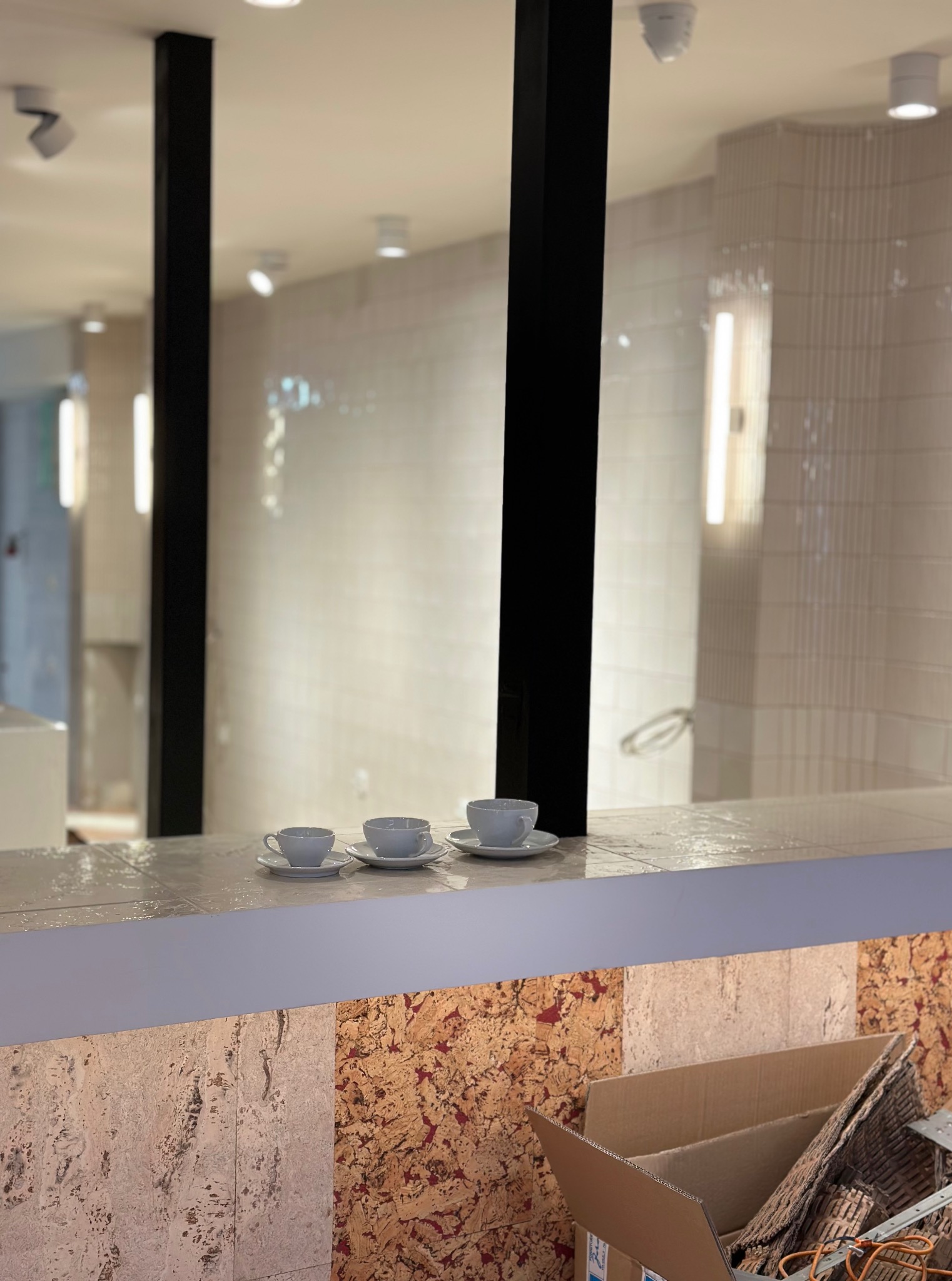 Wykończona łazienka z jasnymi płytkami i trzema filiżankami na blacie. Widoczne detale wykończeniowe oraz elementy dekoracyjne. W tle karton z narzędziami.