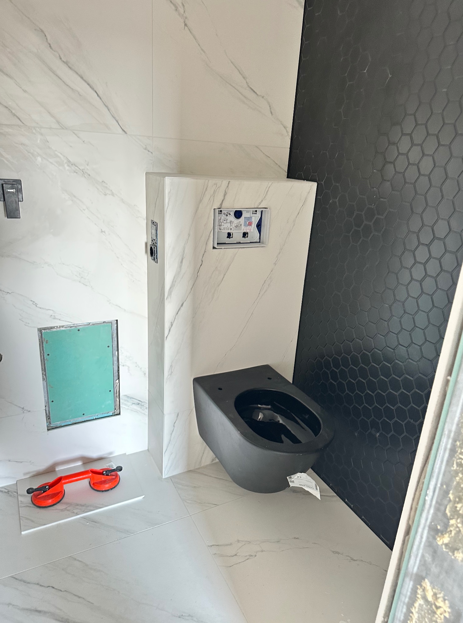 Nowoczesna łazienka w trakcie wykańczania: czarna toaleta podtynkowa, białe płytki imitujące marmur i czarne heksagonalne płytki na ścianie. Widoczne narzędzia glazurnicze.