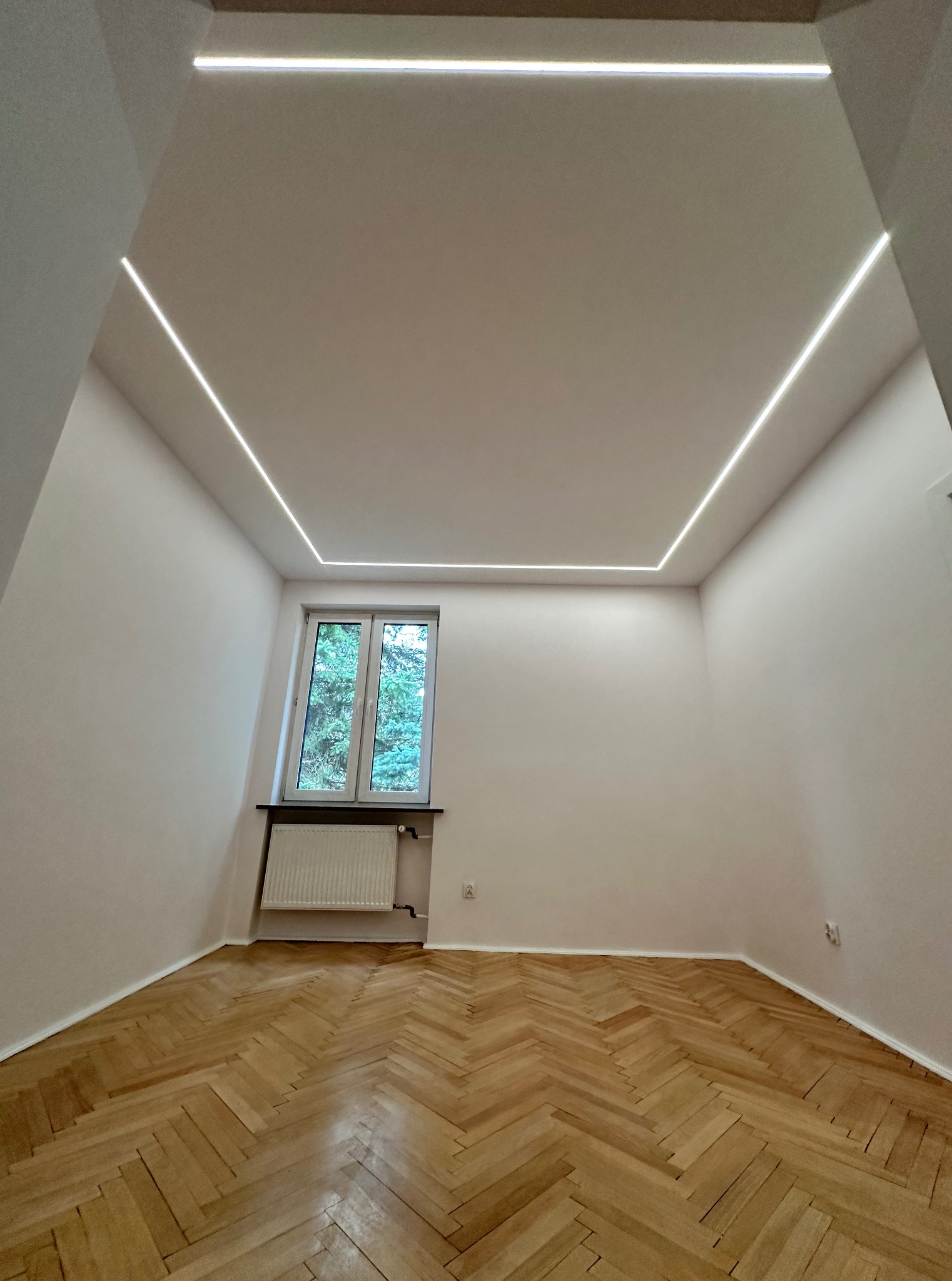Pusty pokój po remoncie: parkiet w jodełkę, okno z widokiem na zieleń i nowoczesne oświetlenie LED w suficie, tworzące geometryczny wzór.