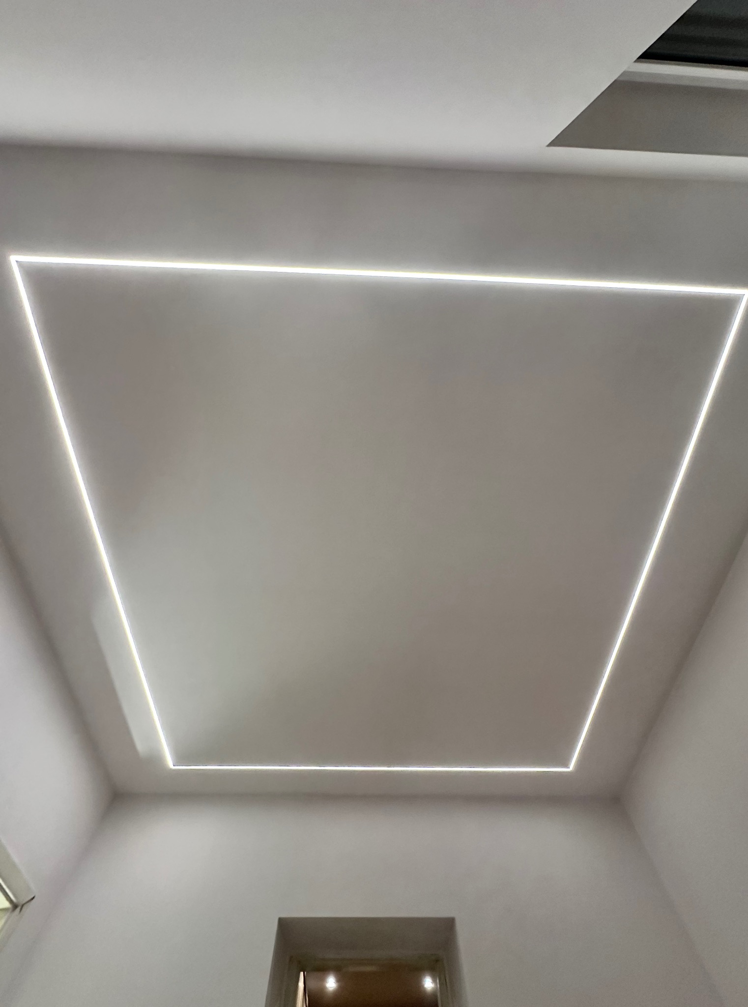 Minimalistyczne oświetlenie sufitowe LED w kształcie kwadratu, w białym pomieszczeniu z widocznym fragmentem okna i drzwi.