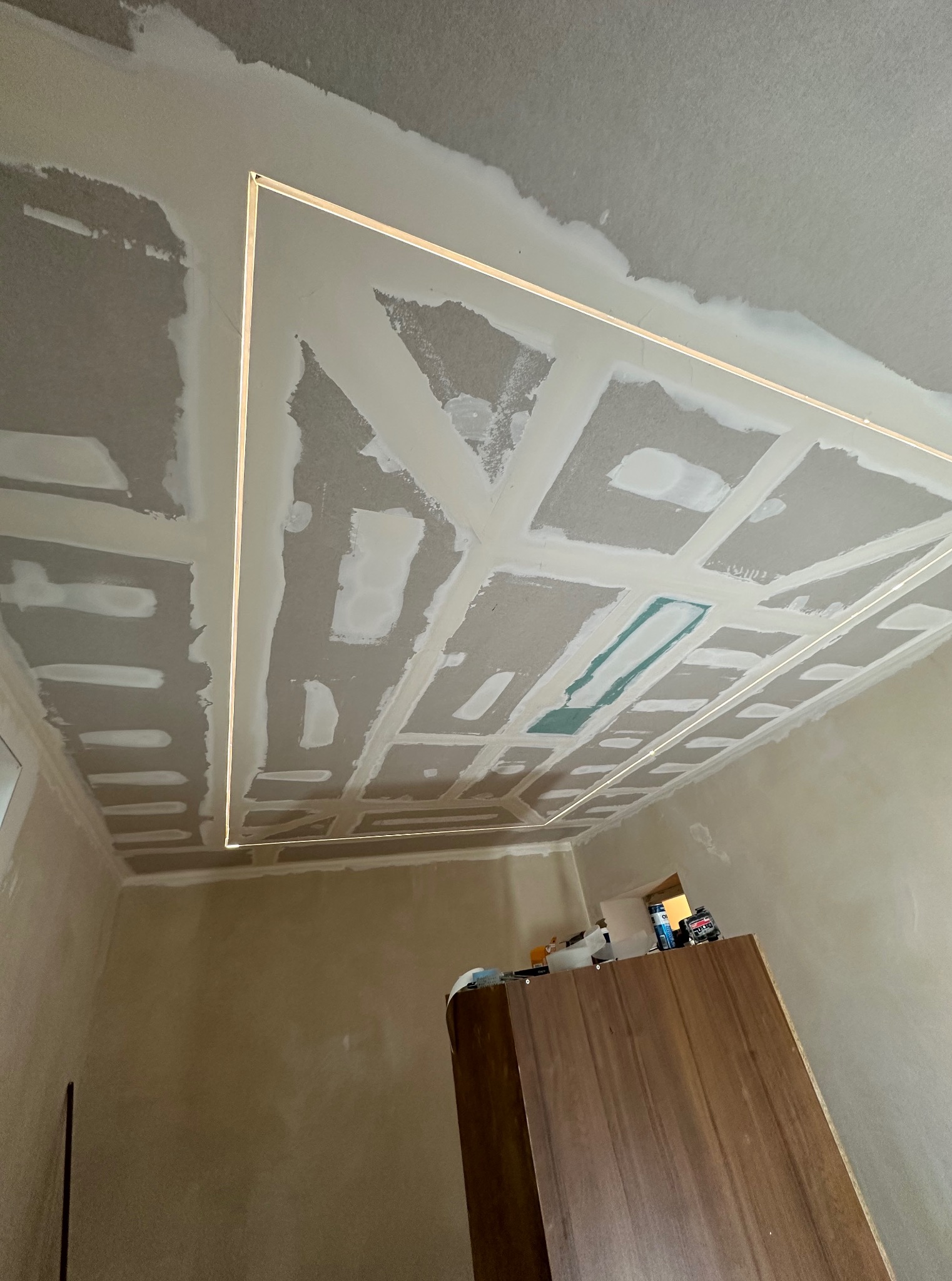 Sufit z płyt kartonowo-gipsowych w trakcie szpachlowania, z widocznym oświetleniem LED wzdłuż krawędzi. Ściany przygotowane do malowania. Widoczny fragment mebla.