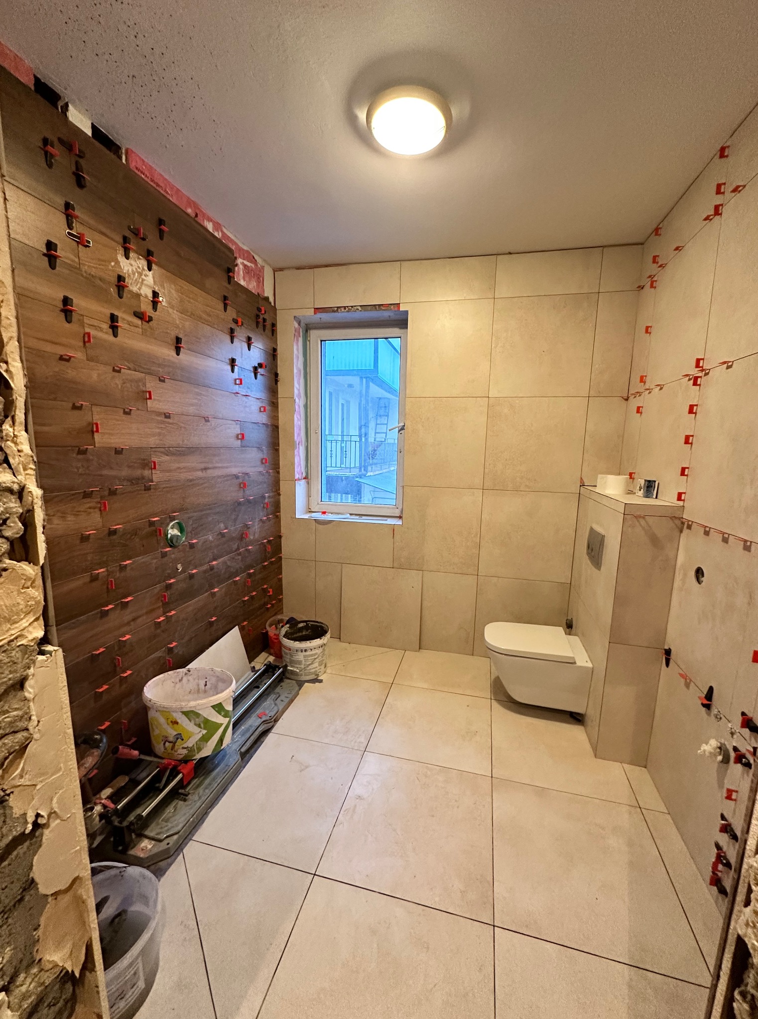 Łazienka w trakcie wykończenia: częściowo ułożone płytki imitujące drewno na ścianie, jasne płytki na podłodze, wisząca toaleta. Widoczne narzędzia glazurnicze i wiaderka.