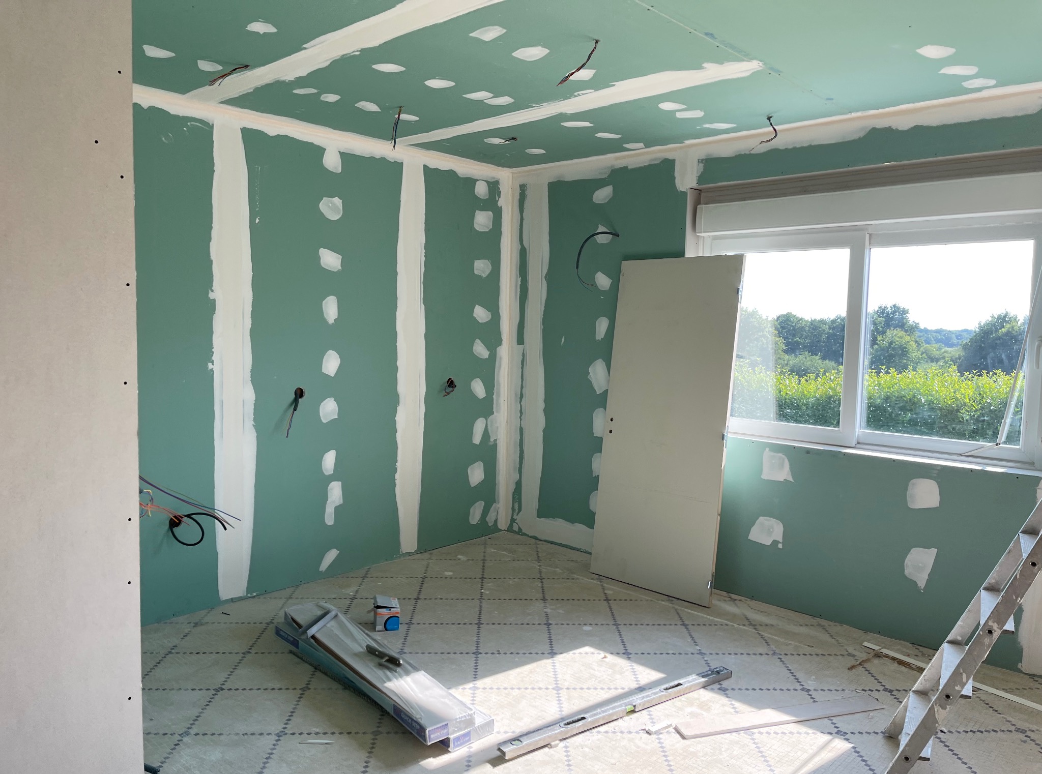 Pokój w trakcie remontu: zielone płyty gipsowo-kartonowe na ścianach, częściowo zaszpachlowane, widoczne przewody, poziomica, drabina. Przygotowanie do dalszych prac.