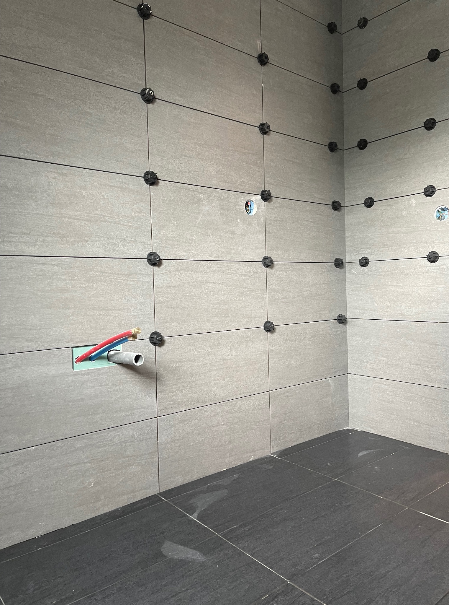 Ściana i podłoga z szarych płytek w trakcie układania, widoczne czarne krzyżyki dystansowe i instalacja wodna. Wykończenie łazienki w trakcie realizacji.