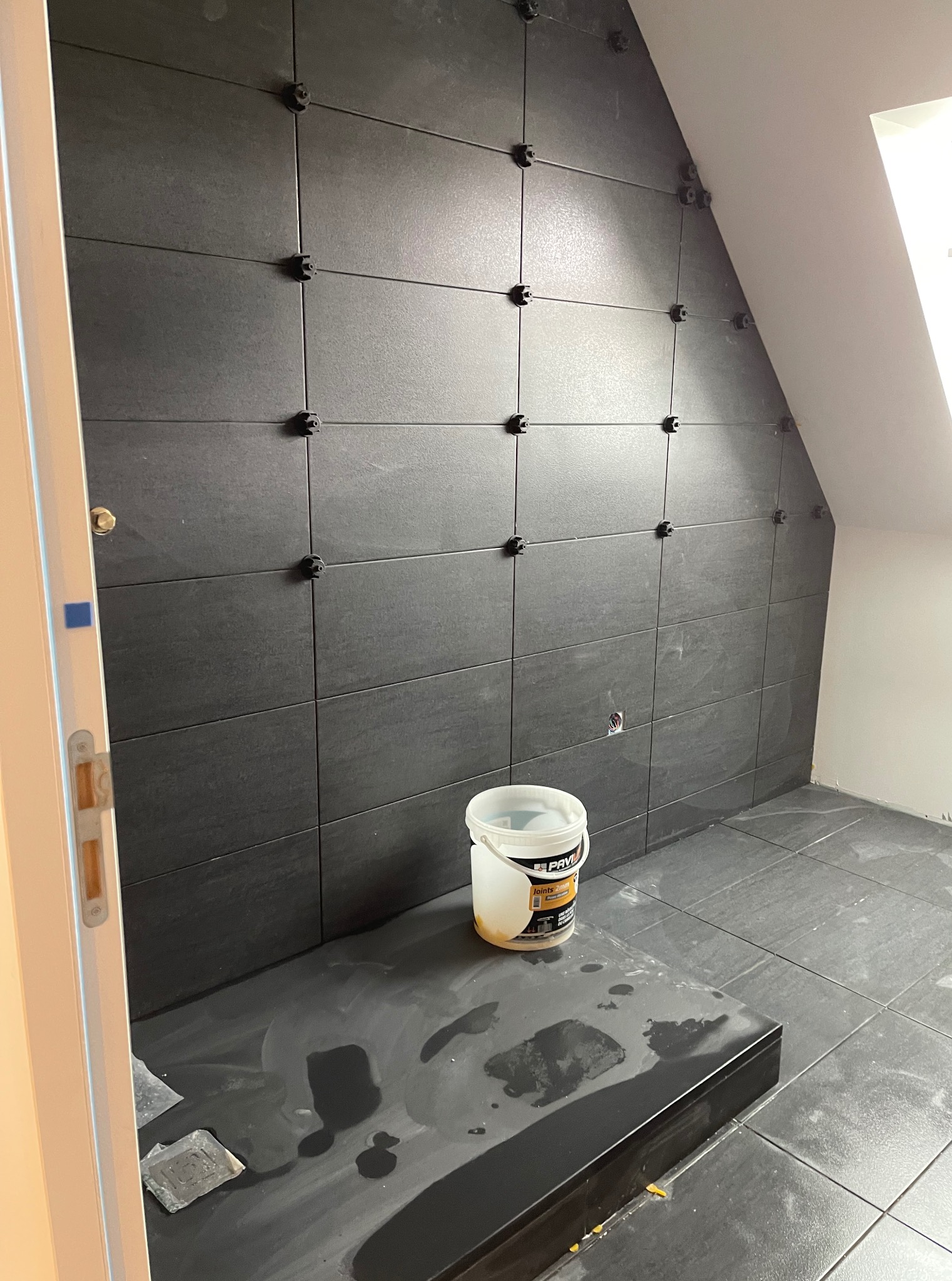 Ciemne płytki na ścianie pod skosem, z czarnymi krzyżykami dystansowymi. Widoczne wiadro z klejem i fragment podłogi. Praca w trakcie wykończenia łazienki.