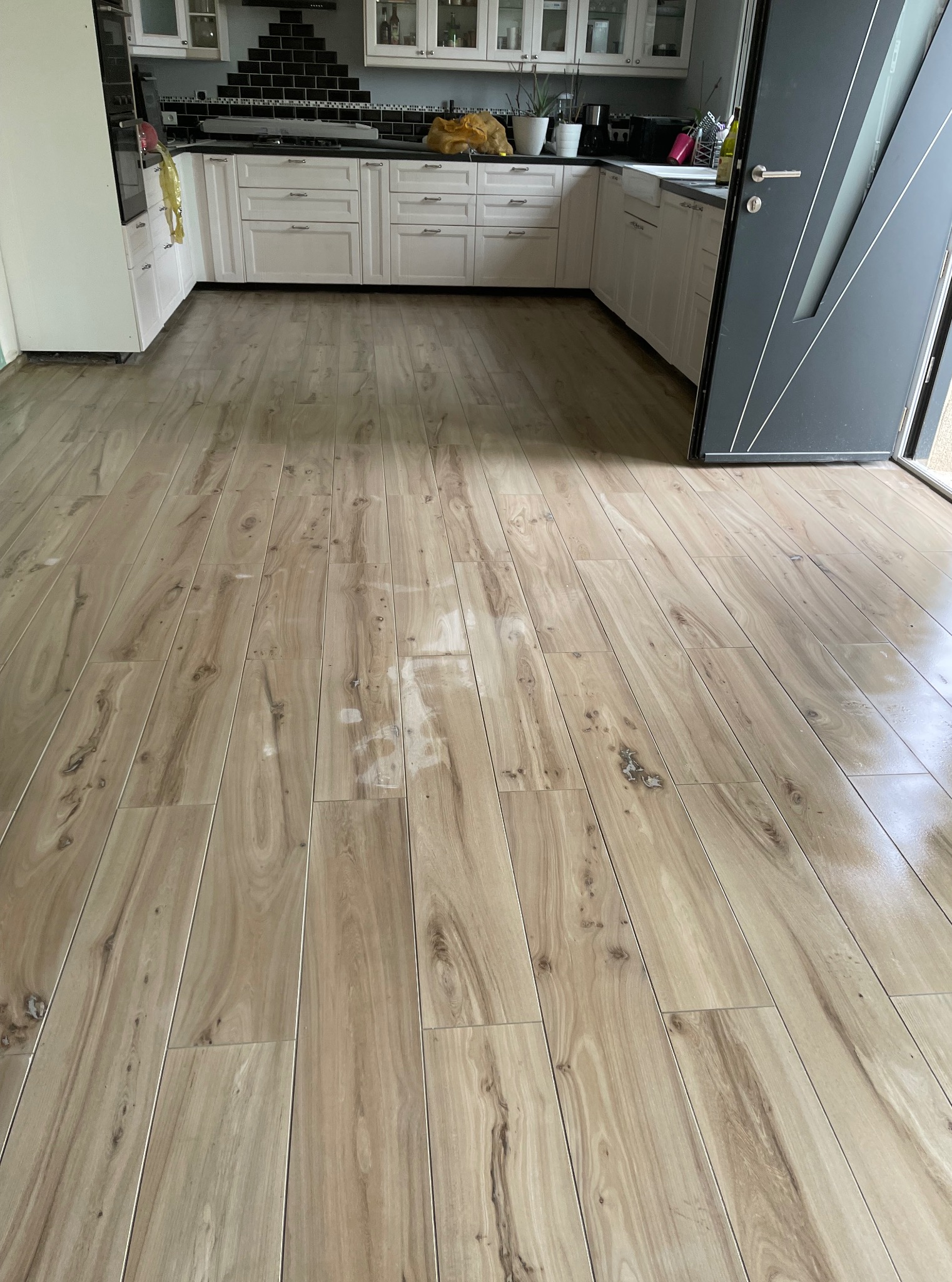 Ułożona podłoga z paneli imitujących drewno w jasnej kuchni z białymi szafkami i czarną mozaiką nad blatem. Widoczna nowoczesna, szara rama drzwi.