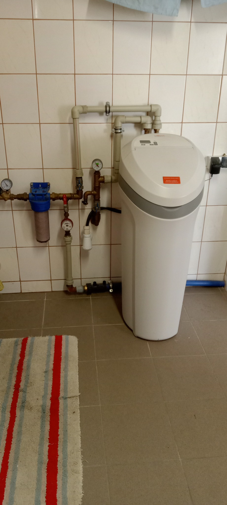 Centralne zmiękczanie wody w kotłowni, widoczne przyłącza hydrauliczne, filtry i manometry na tle ceramicznej ściany.