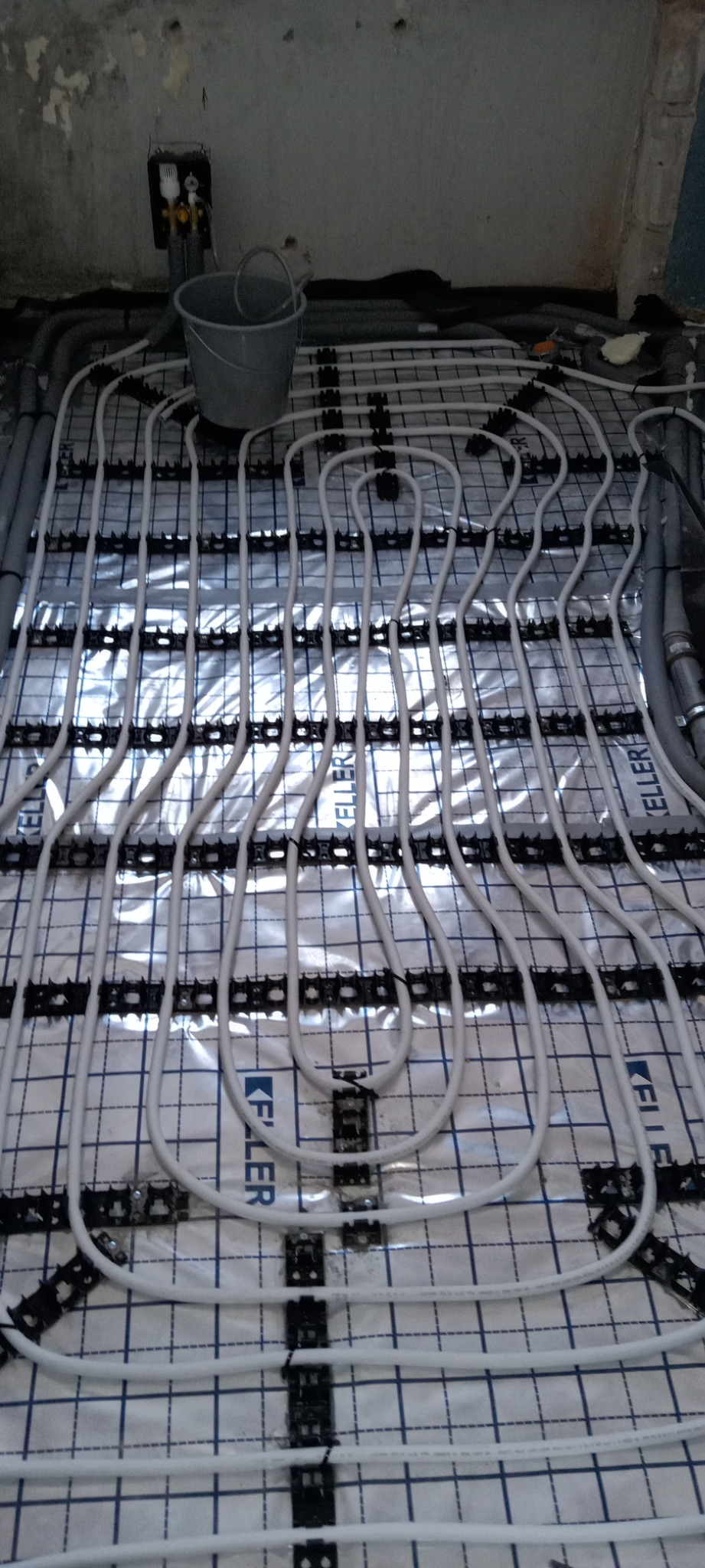 Montaż ogrzewania podłogowego: białe rurki grzewcze ułożone na folii Keller z czarnymi uchwytami mocującymi, widoczny fragment ściany w stanie surowym i wiadro.