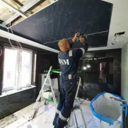 Pracownik w kombinezonie BSM montuje czarny panel sufitowy z wzorem przypominającym marmur, stojąc na aluminiowej drabinie w łazience z marmurowymi ścianami, widoczne metalowe profile konstrukcyjne...