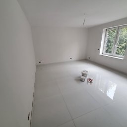 EPP Oskar Kasperkowiak - Pomalowane na biało puste pomieszczenie z błyszczącą podłogą, widoczne puszki z farbą i tubki z masą uszczelniającą, okno z widokiem na zieleń.