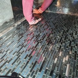 EPP Oskar Kasperkowiak - Układanie mozaiki szklanej w odcieniach szarości i czerni na ścianie, widoczne ręce wykonawcy w różowej bluzie z białymi mankietami.